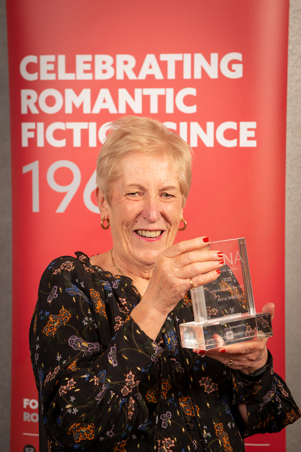 Woman with short blonde hair and patterned black dress holding glass RNA award