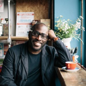 Portrait of Mike Gayle, writer. A black man sits in a cafe wearing a stylish black suit, glasses, coffee cup on a ledge next to him.