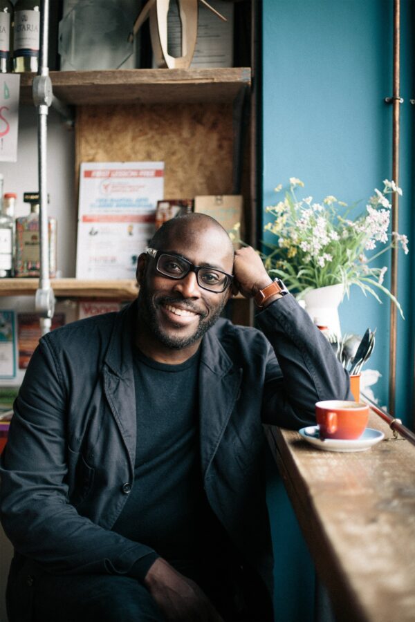 Portrait of Mike Gayle, writer. A black man sits in a cafe wearing a stylish black suit, glasses, coffee cup on a ledge next to him.