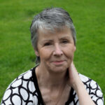 Author headshot of Anni Rose, Short grey hair, black and white top, grass background