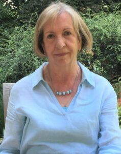 Picture of author Suzanne Merchant. Bobbed blonde hairand light blue collared shirt in front of greenery.