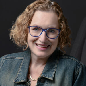 Portrait of Sue Moorcroft, president of the romantic novelist association, curly blonde bob, blue glasses, smiling and jean jacket