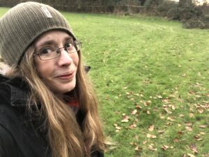 Woman with long blonde hair, glasses and hat outside in green space