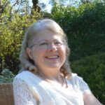 Author Linda Corbett sitting in garden wearing white shirt, glasses, and smiling
