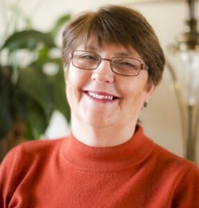 A photograph of Elaine Roberts. She has short brown hair, glasses, a jumper and a smile.
