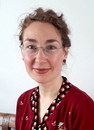 A photo of Ruth. She is smiling, she's wearing glasses and a bright red cardigan.