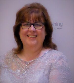 Author Audrey Harrison, dark shoulder length hair, black glasses, smiling, white shirt with lace at top