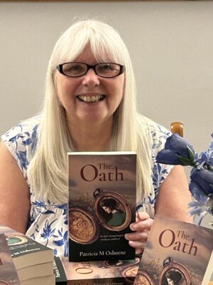 Writer Tricia Osborne, Blonde hair, black glasses, holding a copy of her book the oath